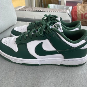Nike Dunk Low vit/grön sneakers - Nike Dunk Low sneakers i vitt och mörkgrönt skinn med klassisk swoosh på sidorna. Lågt skaft, gröna snören och vit/green yttersula. Perfekt för dig som gillar streetstyle och vill sticka ut med en ikonisk modell.