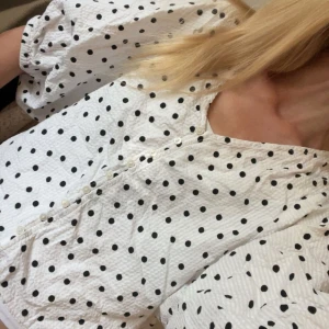 Prickig blus med puffärm från Lindex - Superfin vit blus från Lindex med svarta prickar, v-ringning och knappar hela vägen framtill. Blusen har puffiga korta ärmar och är gjord i ett strukturerat bomullstyg som känns både luftigt och trendigt. Perfekt för dig som gillar lekfulla mönster och snygga detaljer.