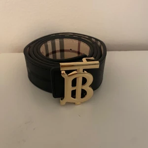 Burberry bälte med guldspänne - Snyggt Burberry bälte med klassiskt rutigt mönster i beige, svart och vinrött. Bältet har ett stort guldigt 'B'-spänne och är tillverkat i skinn. Insidan har Burberrys ikoniska rutor och utsidan är svart med subtilt mönster. Perfekt accessoar för att lyfta din outfit.