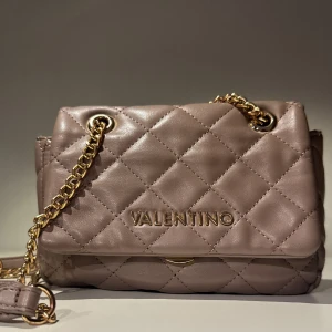 Quiltad axelväska från Valentino - Säljer en snygg quiltad axelväska från Valentino i beige skinnimitation med guldfärgad kedja. Väskan har ett klassiskt rutmönster, magnetlås och flera fack inuti, inklusive ett med dragkedja. Perfekt storlek för det viktigaste och ger en lyxig känsla.