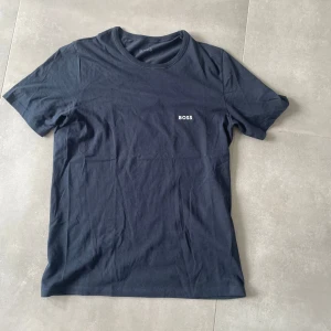 Mörkblå t-shirt från BOSS - Snygg mörkblå t-shirt från BOSS med klassisk passform och rund hals. T-shirten har en stilren design med diskret BOSS-logga broderad på bröstet. Tillverkad i mjuk bomull som känns skön mot huden. Perfekt för en clean och enkel look.
