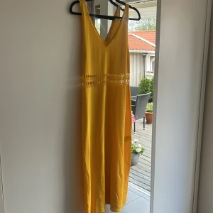 Gul långklänning med spetsdetalj - Säljer en solgul långklänning i medium med snygg v-ringning och ärmlös design. Klänningen har en bred spetsdetalj runt midjan som ger extra stilpoäng. Materialet är mjukt och lätt, perfekt för varma dagar. Passar dig som gillar färg och vill sticka ut.