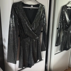 Glittrig silver kortklänning H&M - Säljer en glittrig silverfärgad jumpsuit från H&M med omlott-design och djup v-ringning. Den har långa ärmar och markerad midja, perfekt för att sticka ut. Materialet är syntet med paljett-detaljer som ger extra shine.