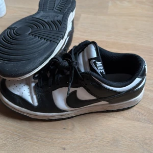 Nike Dunk Low svart/vit sneakers - Nike Dunk Low sneakers i svart och vitt med klassisk design. Skorna har svart snörning, vit tåbox och svarta detaljer längs sidorna samt vit Nike-logga på hälen. Ovandelen är i skinn och insidan är fodrad med svart textil. Perfekta för dig som gillar streetstyle.