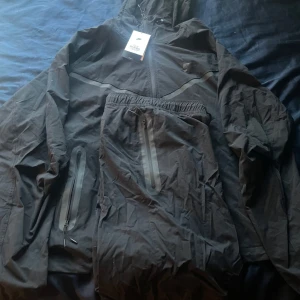 Svart vindjacka från Nike med huva - Svart vindjacka från Nike med huva och dragkedja framtill. Jackan har en loose fit och reflekterande detaljer över bröstet. Praktiska fickor med dragkedja och lätt material gör den perfekt för aktiva dagar. Snygg och sportig stil med diskret Nike-logga på bröstet.