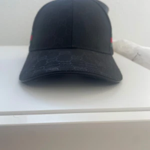 Svart keps med ett coolt mönster - Snygg svart keps  med diskret monogram över hela kepsen och klassiska gröna och röda ränder på sidan. Justerbar rem bak i svart skinn med präglad.