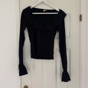 Svart långärmad topp med volang - Supersnygg svart topp från nelly med långa ärmar och bred volang runt ringningen. Ärmarna har extra volang vid ärmsluten för en trendig look. Perfekt för dig som gillar detaljer och vill sticka ut lite extra.