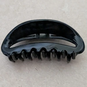 Svart hårklämma i plast - En enkel och stilren svart hårklämma med rundad form och starkt grepp. Perfekt för att snabbt sätta upp håret och hålla det på plats. Klämman har en blank finish och är tillverkad i tålig plast med klassisk design.