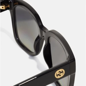 Svarta fyrkantiga solglasögon från Gucci - Snygga svarta solglasögon från Gucci med fyrkantig form och breda skalmar. Glasen är mörka och har en snygg toning. På sidan sitter den klassiska Gucci-loggan i guld. Ramen är tillverkad i blank syntet och ger en cool vibe. Helt äkta och använt endast 1 gång tidigar. 