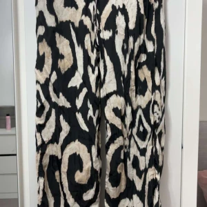 Svarta vida byxor med mönster H&M XS - Säljer ett par riktigt snygga vida byxor från H&M i storlek XS. Byxorna har ett svart och beige abstrakt mönster, resår i midjan och är tillverkade i ett lätt och luftigt material. Perfekta för dig som vill sticka ut med en cool look.
