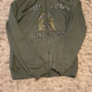 Grön zipjacka från Blinglucid XS - Snygg grön zipjacka från Blinglucid med blingade detaljer och texten 'CHASE THE BAG' samt 'BLING LUCID' framtill. Jackan har ett stort motiv med pengasäck i guld och silver, dragkedja och långa ärmar. Perfekt för dig som gillar streetstyle och statement-plagg.