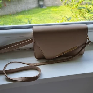 Beige väska med axelrem - Snygg beige kuvertväska med lång axelrem och stilren guldfärgad detalj på framsidan. Väskan har ett minimalistiskt uttryck och är tillverkad i slätt syntetmaterial. Perfekt storlek för mobil och småsaker, med svart foder invändigt.