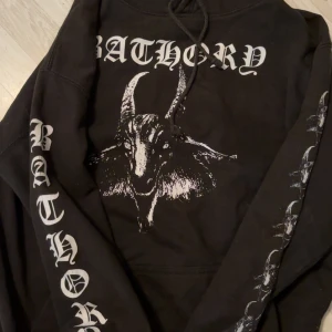Bathory svart hoodie med tryck - Svart hoodie från Bathory med stort getmotiv och logga på bröstet, tryck längs ärmarna och stor pentagramgrafik på ryggen. Klassisk huva med snörning och känguruficka framtill. Perfekt för dig som gillar metal och vill sticka ut.