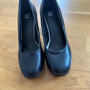 Svarta klackar  - Snygga svarta pumps från XIT i storlek 39. Skorna har en rund tå och hög klack med plattform, vilket ger extra höjd och stabilitet. Tillverkade i syntetmaterial med en stilren och klassisk design som passar till många olika outfits.