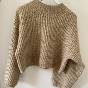 Beige stickad croppad tröja - Säljer en beige stickad tröja med croppad passform och breda, långa ärmar. Tröjan har en ribbad struktur och en hög, ribbad halsringning som ger en chill och trendig vibe. 