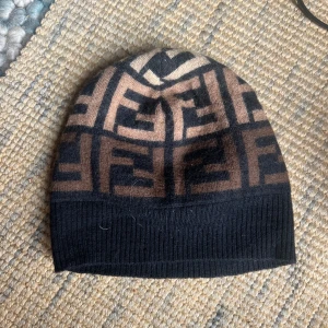 Fendi FF-mönstrad mössa i ull - Snygg mössa från Fendi med ikoniskt FF-mönster i svart och brunt. Tillverkad i mjuk ull och har ribbad kant nedtill för extra komfort. Perfekt accessoar för att lyfta din look med lyxig streetstyle-vibe. Äkta från vestaire! Säljer för den inte är min stil och jag vill mest ha bort den.