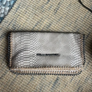 Clutch  - Kuvertväska i beige med ormskinnsmönster och snygga kedjedetaljer längs kanterna. Väskan har dragkedjefack på insidan och stängs med tryckknappar. Väskan är inte äkta