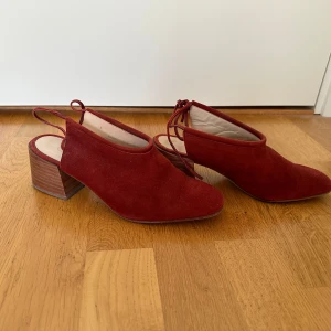 Vinröda mocka klackskor från COS - Snygga vinröda klackskor från COS i mjuk mocka. Skorna har fyrkantig tå, öppen häl med smal rem och knytning baktill. Klacken är bred och stabil i trälook, vilket ger en cool och modern vibe. Perfekta för dig som vill sticka ut med stilrena detaljer.