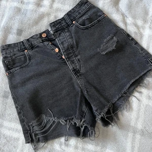 Svarta jeansshorts från Zara - Snygga svarta jeansshorts från Zara med hög midja och råa, fransiga kanter. Shortsen har slitna detaljer framtill och klassisk femficksdesign. Perfekta för en chill sommarlook med lite edge.