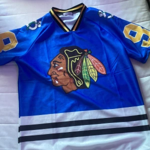 Blå Bedard hockeytröja #98 - Säljer en blå hockeytröja med Chicago Blackhawks-logga på bröstet och stora gula siffror 98 samt namnet Bedard på ryggen. Tröjan har svarta och vita ränder nertill och detaljer i gult och svart vid kragen. Perfekt för hockeyfans!