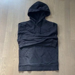 Svart hoodie med huva och magficka - En klassisk svart hoodie med huva och dragsnören. Tröjan har en stor magficka framtill och ribbade muddar vid ärmslut och nederkant. Perfekt för en chill och avslappnad stil. Materialet känns mjukt och skönt mot huden.