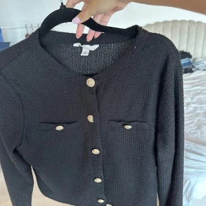 Svart kofta med guldknappar  - Svart kofta från H&M i storlek M med snygg struktur och rund hals. Koftan har dekorativa guldknappar framtill och på de två bröstfickorna. Långärmad och tillverkad i ett stickat material som känns mjukt och lite lyxigt. Använd fåtal gånger med inga slitage eller defekter.