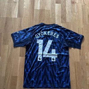 Arsenal bortatröja Gyökeres #14 - Snygg Arsenal bortatröja från Adidas med Gyökeres och nummer 14 på ryggen. Tröjan har ett elektriskt blått och svart blixtmönster, röda Adidas-ränder på axlarna och tryckta sponsorer. Tillverkad i lätt och ventilerande polyester, perfekt för match eller träning.