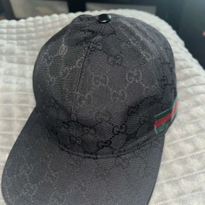 Svart Gucci keps med GG-mönster - Snygg svart keps från Gucci med klassiskt GG-monogram över hela kepsen. På sidan finns den ikoniska gröna och röda randen. Justerbar rem baktill och böjd skärm. Perfekt accessoar för att lyfta din streetwear-look.