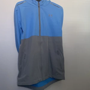 Blå och grå jacka från Under Armour - Snygg blå och grå jacka från Under Armour med dragkedja och huva. Perfekt för träning eller en avslappnad dag. Jackan har praktiska fickor med dragkedja framtill. Xs men passar S för den är lite oversized 