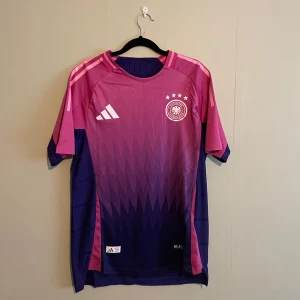 Tyskland fotbollströja från Adidas - Snygg fotbollströja från Adidas i rosa och lila med korta ärmar. Tröjan har ett coolt mönster och det tyska fotbollsförbundets emblem på bröstet. Perfekt för fotbollsfans som vill sticka ut på planen!