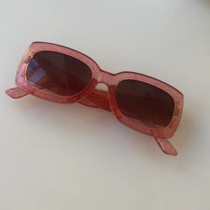 Glittriga rosa solglasögon - Snygga rektangulära solglasögon i en glittrig rosa färg. Perfekta för att ge din outfit en extra touch av glamour. De har en bred båge och mörka linser som skyddar mot solen.