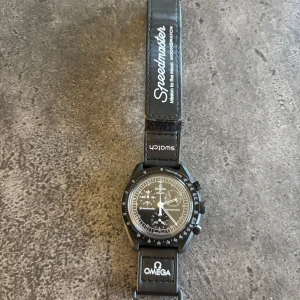 Svart klocka från Omega x Swatch moonphase - Säljer en stilren svart klocka från Omega x Swatch. Klockan har en klassisk design med vit text och detaljer på urtavlan. Armbandet är svart med Omega och Swatch-logotyper. Perfekt för den som gillar tidlös design. Fick denna i present så har inget kvitto och hittar tyvärr inte boxen därav priset