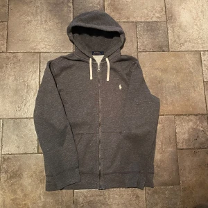 Grå Zip Hoodie från Ralph Lauren - Säljer en stilren grå Zip hoodie från Ralph Lauren med dragkedja och klassisk logga på bröstet. Tröjan har långa ärmar och en justerbar huva med snören. Perfekt för en avslappnad look. Extremt gott skick.