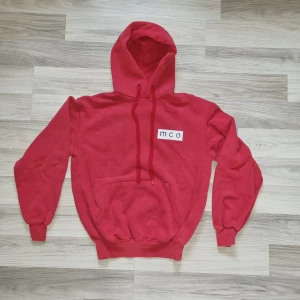 Röd hoodie från Fruit of the Loom - Säljer en klassisk röd hoodie från Fruit of the Loom med en vit mco-logga på bröstet. Den har en stor ficka framtill och justerbar huva med dragsko. Perfekt för en avslappnad stil. 🧢