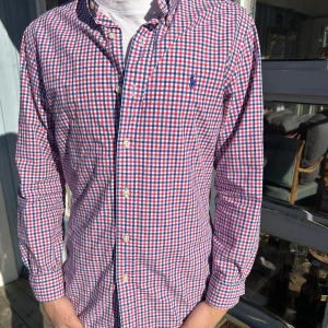 Rutig skjorta från Ralph Lauren - Säljer en stilren rutig skjorta från Ralph Lauren i blått, rött och vitt. Skjortan har en slim fit passform och är långärmad med knappar framtill. Perfekt för en snygg och avslappnad look. Knappen är lite söndrig på vänstra armen men märks inte!
