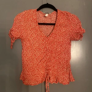 Röd blommig blus från H&M - Charmig röd blommig blus från H&M med korta ärmar och v-ringning. Blusen har knytband framtill och volangdetaljer vid ärmslut och nederkant. Perfekt för en somrig look! 🌸