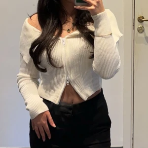 Vit ribbad offshoulder kofta - Säljer en stilren vit ribbad kofta med offshoulder-design och dragkedja framtill. Perfekt för en trendig look.