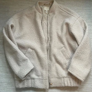 Beige teddyjacka från H&M - Mysig beige teddyjacka från H&M med dragkedja framtill. Jackan har en avslappnad passform och är perfekt för kyligare dagar. Den har långa ärmar och en mjuk, fluffig yta som ger en trendig look.
