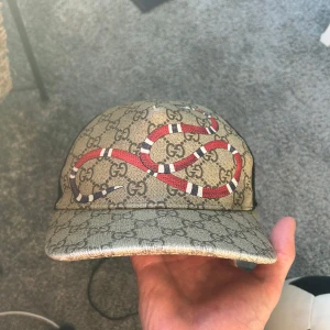 Beige keps med ormmönster från Gucci - Snygg beige keps från Gucci med ett coolt ormmönster i rött och svart. Kepsen har det klassiska GG-mönstret och justerbar rem baktill med röd och grön rand. Perfekt för att ge din outfit en lyxig touch.