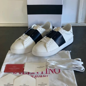 Valentino Garavani skor - Snygga Valentino Garavani sneakers i vitt med en bred svart rem över ovansidan. Skorna har en klassisk design med snörning och en bekväm sula. Perfekta för en stilren look.