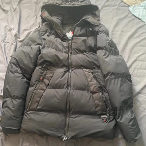 Svart dunjacka från Zavetti Canada - Säljer en svart dunjacka från Zavetti Canada med huva och dragkedja. Jackan har en broderad lönnlövdetalj på ärmen och är perfekt för kalla dagar. Den har praktiska fickor med dragkedjor och en stilren design. Storlek xs men passar s