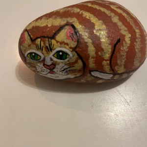 Handmålad sten med kattmotiv - Unik handmålad sten föreställande en söt katt med gröna ögon. Stenen har en brun och gulrandig design med detaljerade ansiktsdrag. Perfekt som dekoration eller present till kattälskare.