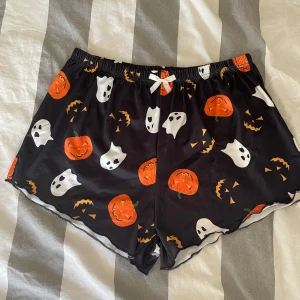 Svarta shorts med Halloween-motiv - Säljer ett par svarta shorts med ett coolt Halloween-motiv av pumpor och spöken. De har en elastisk midja med en söt vit rosett framtill. Perfekta för en avslappnad och rolig stil!