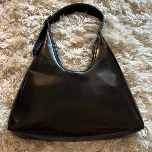 Svart axelväska i syntet - Snygg svart axelväska i glansigt syntetmaterial. Väskan har en enkel och stilren design med en bred axelrem som gör den bekväm att bära. Från Carin Wester och inköpt sommaren 2024.