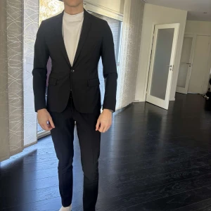 Svart kavaj från Dondup - Snygg svart kavaj från Dondup med en klassisk design. Kavajen har en enkelknäppning och smal passform, perfekt för en stilren look. Den är tillverkad i ett elegant material och har en ficka med märket på insidan. Byxor finns att köpa till för 200 kr om man köper kavajen. 