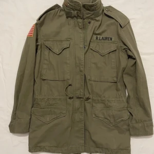 Ralph lauren field jacket - Säljer en olivgrön jacka från Ralph Lauren med broderad amerikansk flagga på ärmen och flera praktiska fickor. Jackan har en hög krage och dragkedja framtill. Perfekt för en cool och avslappnad stil.