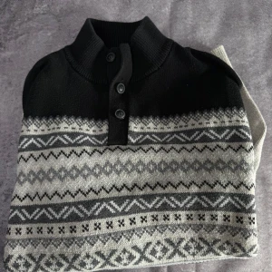 Svart och grå mönstrad tröja från Chaps - Säljer en snygg svart och grå tröja från Chaps med ett stilrent mönster. Tröjan har långa ärmar och knappar vid halsen. Perfekt för kyligare dagar.