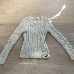 Vit stickad tröja från Hollister - Säljer en snygg vit stickad tröja från Hollister med kabelstickat mönster. Tröjan har långa ärmar och en liten röd logga broderad längst ner. Perfekt för kyliga dagar! 🧶