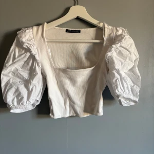 Vit topp från Zara - Säljer en söt vit blus från Zara med puffiga korta ärmar och en fyrkantig urringning. Blusen har en ribbad struktur och elastiska ärmslut. Använd 1/2 gånger, strl S men funkar som xs