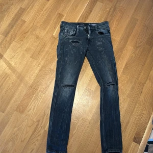 Svarta jeans från Replay - Snygga svarta jeans från Replay, modell Anbass. De har en slim fit och är dekorerade med slitningar på knäna och låren för en trendig look. Perfekta för dig som vill ha en stilren men ändå edgy stil. 
